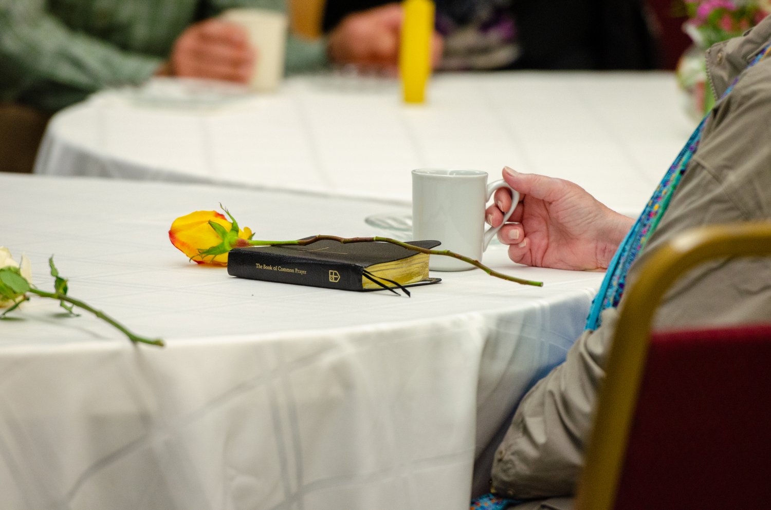 Person's hand holding coffee at table beside a prayer book and a flower