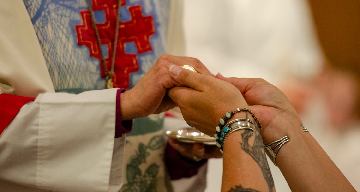 Closeup of two hands clasped, one person wearing vestments, the other with tattooed wrists