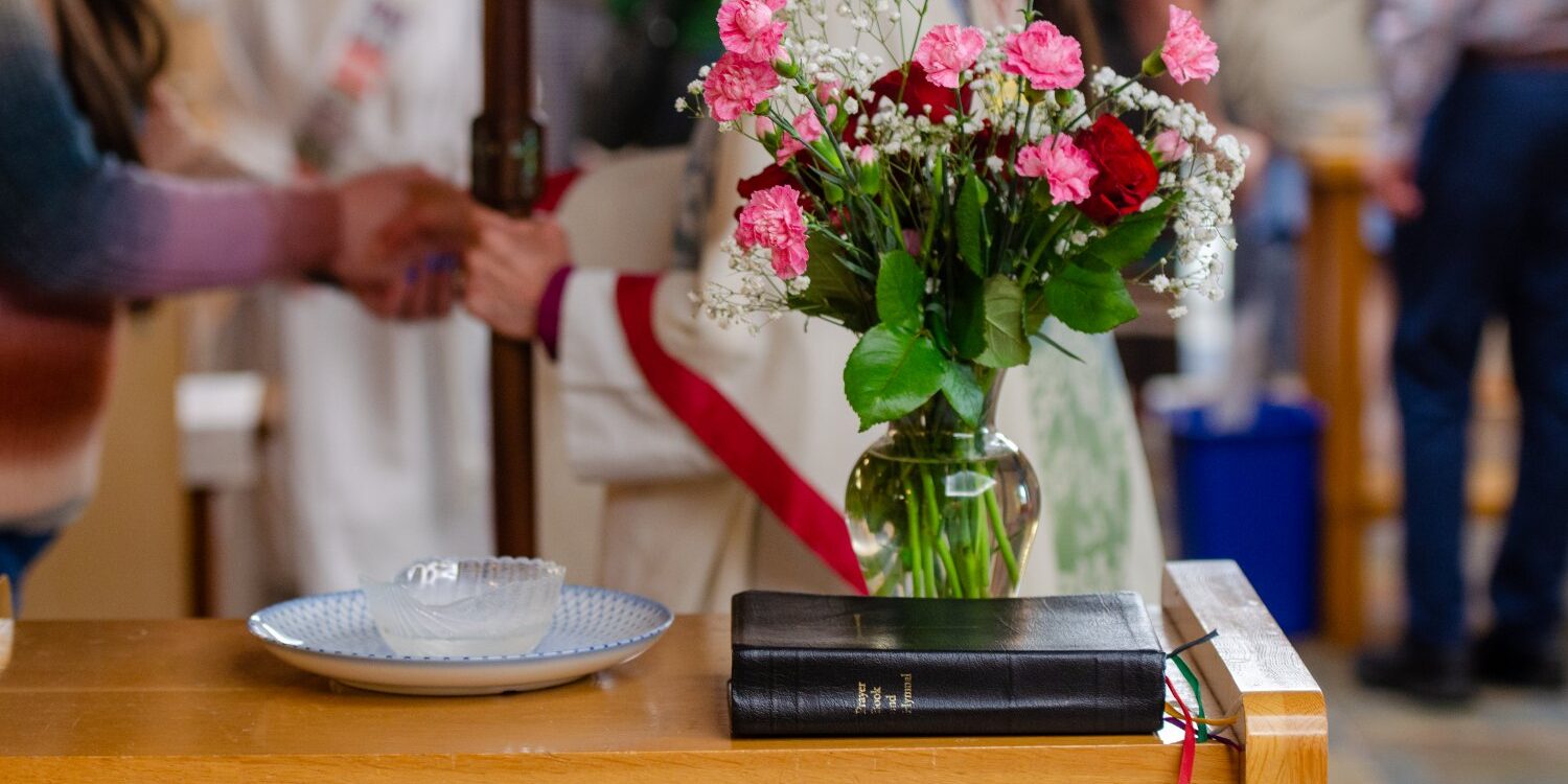 Vase of flowers beside a Book of Common Prayer while people in robes interact in the background