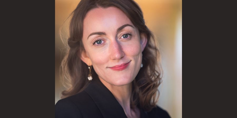 Portrait of a smiling woman with brown hair and blue eyes, wearing a black blazer and earrings.